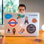 man peers over a computer screen with popular brand stickers on the back